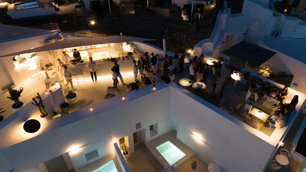 An aerial of the lit cave bar at night, guests gathered before it and two plunge pools glowing turquoise below.