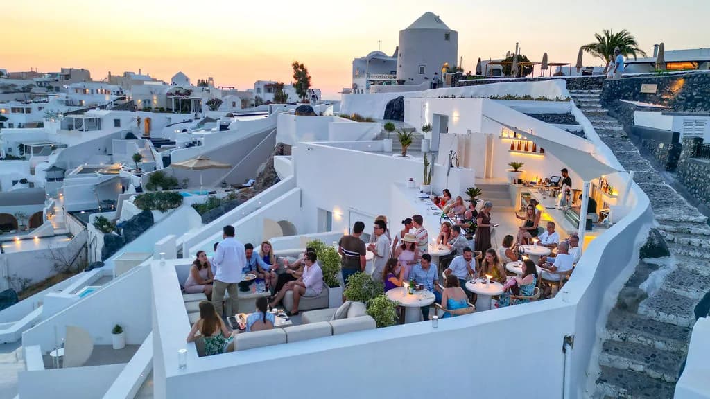 An aerial over the full rooftop at sunset, candlelit tables and sofas below Oia's windmill and the stone steps.