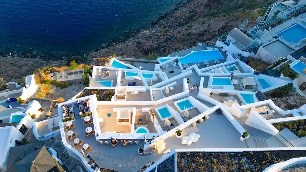 An aerial of whitewashed suites, tables and turquoise plunge pools stepping down the cliff above the deep-blue sea in late-afternoon light.