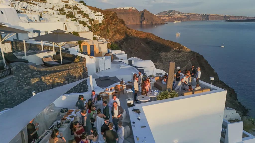 An aerial of a private celebration filling the rooftop in golden light, the caldera cliffs, open sea and anchored boats beyond.