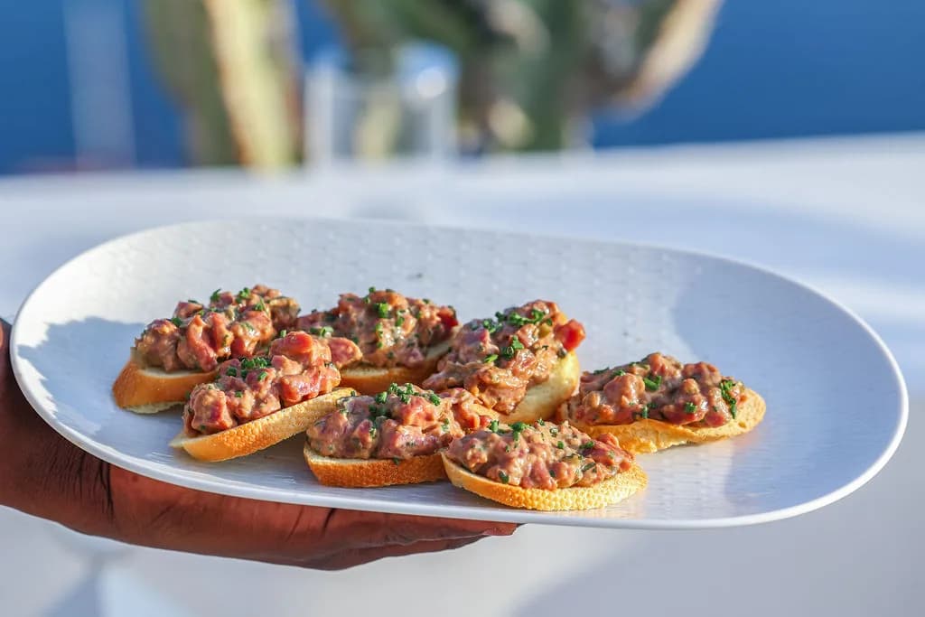 A plate of tartare crostini held out to the table, the sea soft behind it.