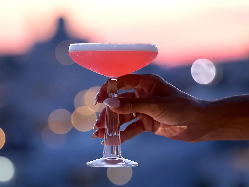 A guest's hand holding a foamed pink coupe by its fluted stem, the sky pink at dusk and the village lights blurred behind.