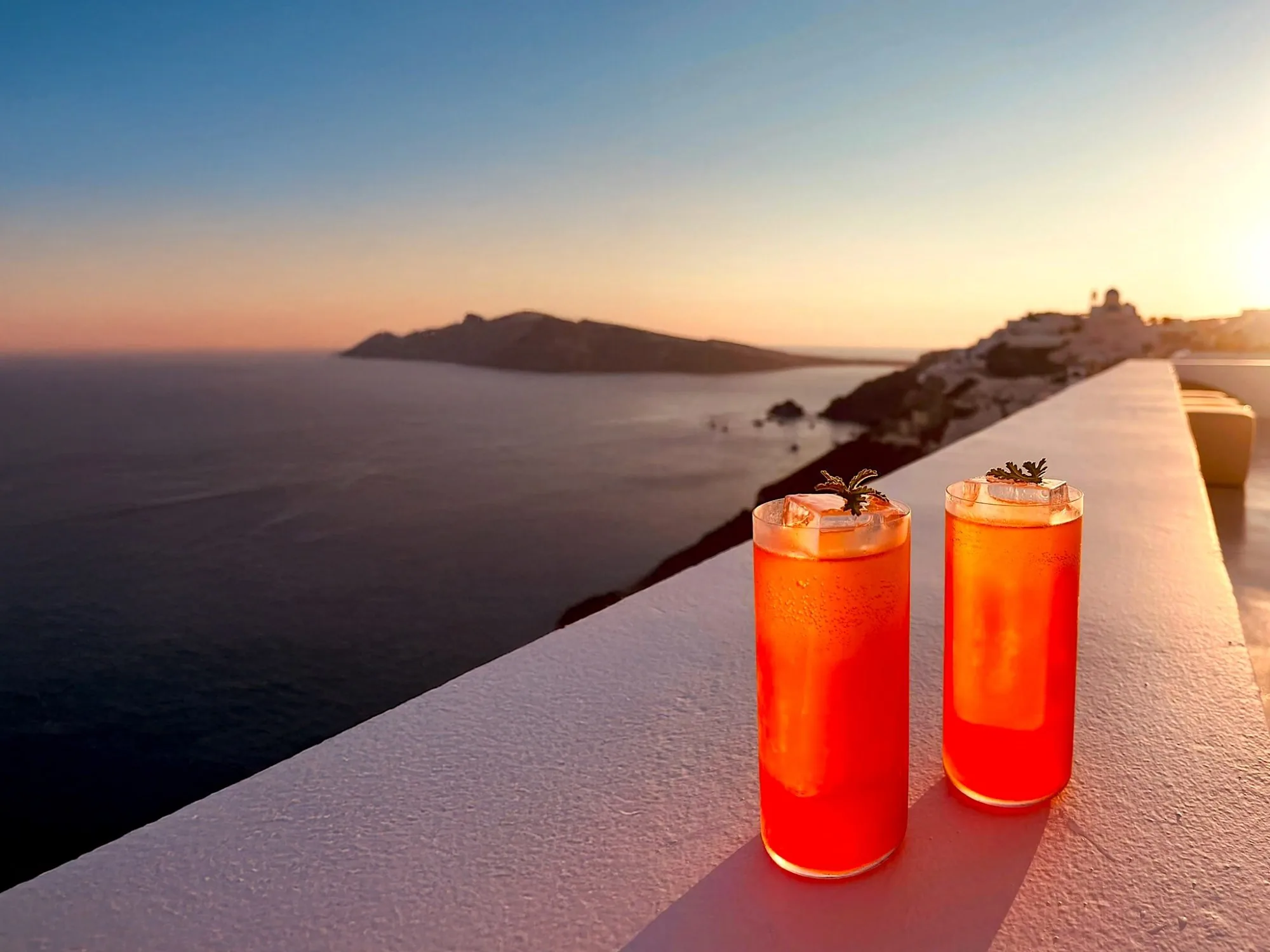 Two orange-red highballs with herb sprigs on the white ledge, the sun setting beyond Oia above the open caldera.