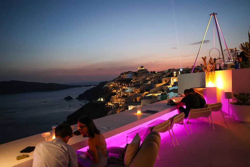 Two guests at the magenta-lit ledge at blue hour, the tripod above them and Oia's lights stepping down to the caldera.