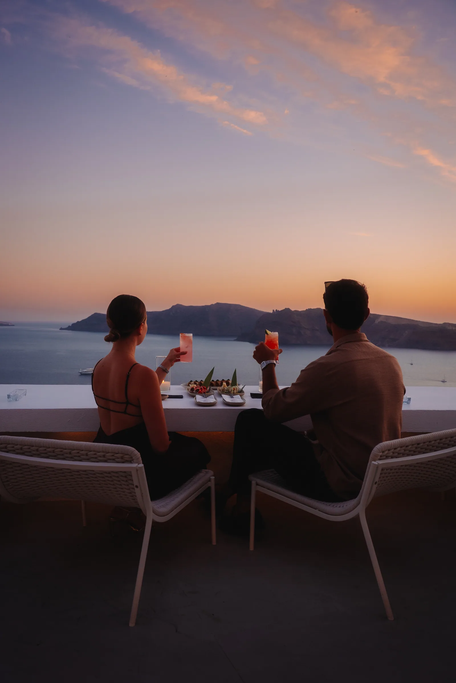 Two guests raise their cocktails at the rooftop ledge as the sky glows over the caldera.