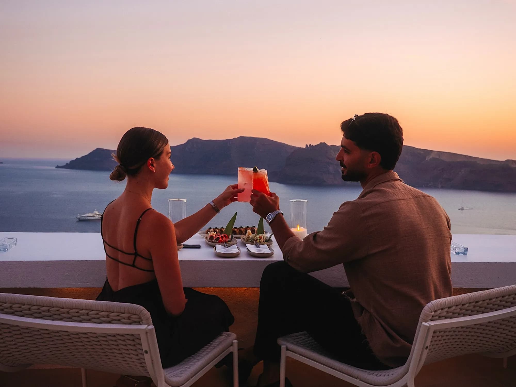 A couple, seen from behind, touching cocktail glasses at the ledge as the sun sets over the caldera, sushi plates and a candle between them.