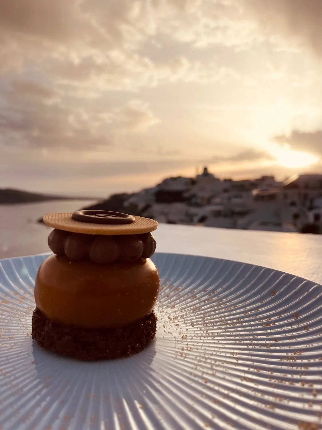 A caramel dessert on a fluted plate, the village and sea behind.