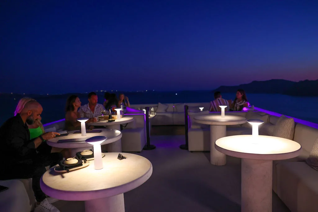 Guests on low white sofas around glowing round tables at blue hour, the sea and the dark caldera rim behind them.