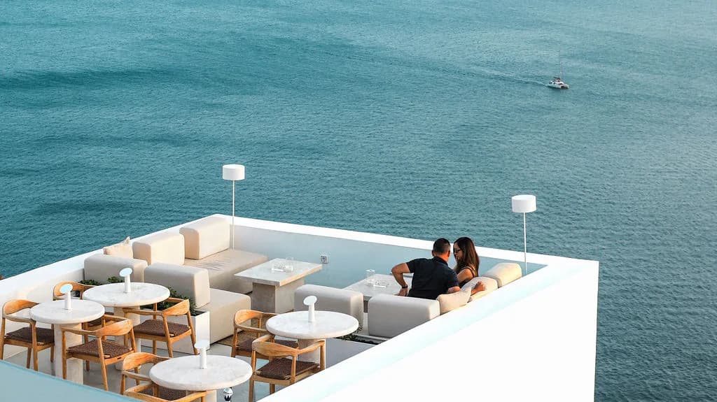 Two guests on a white sofa at the corner of the rooftop, empty tables beside them and a lone boat on the teal sea below.