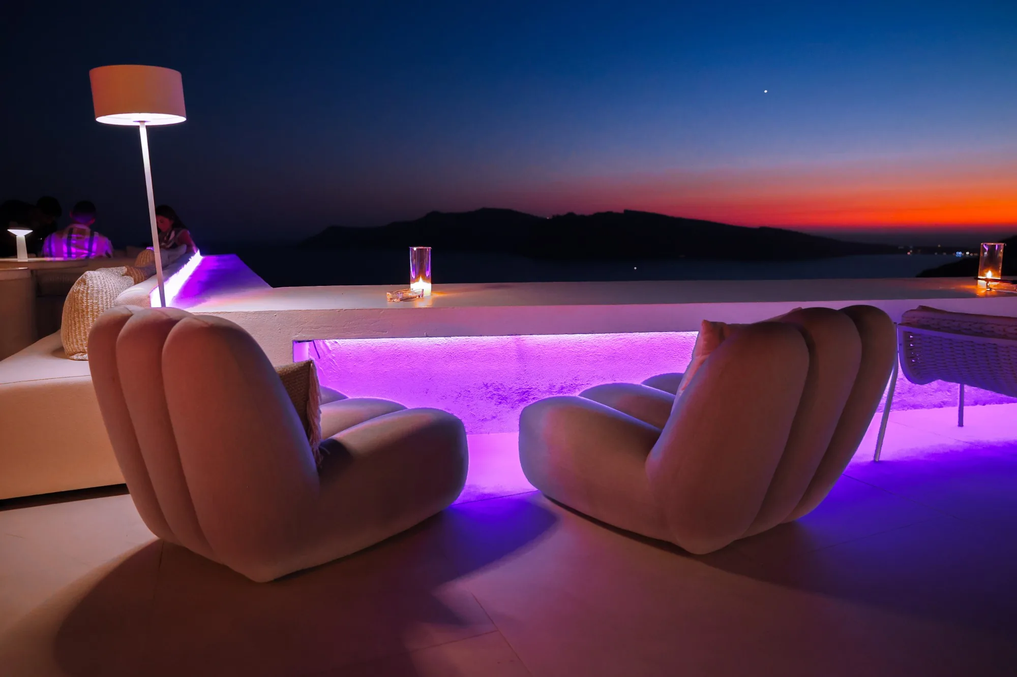 Two curved lounge chairs facing a violet-lit ledge after sunset, a floor lamp and a candle on the ledge, the caldera fading to orange beyond.