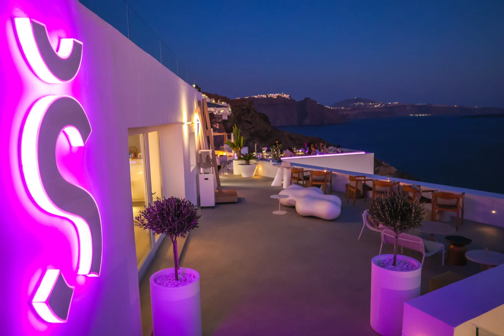 A magenta neon S glowing on the white wall beside a quiet rooftop lounge, the caldera deep blue beyond.
