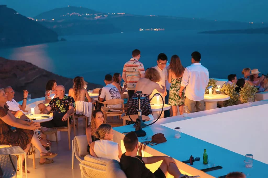 Guests filling the rooftop at blue hour around lit tables and a shisha, the caldera and the lights of the far rim beyond.