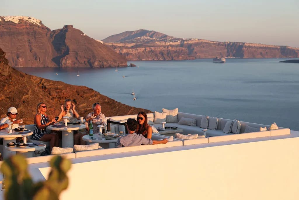 Guests on the long white sofa and at round tables in low golden sun, the caldera cliffs, sailboats and a cruise ship behind.