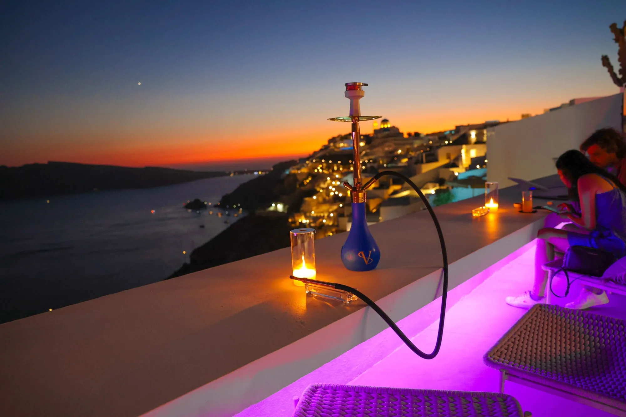 A shisha on the rooftop ledge against the afterglow, Oia lit along the cliff.