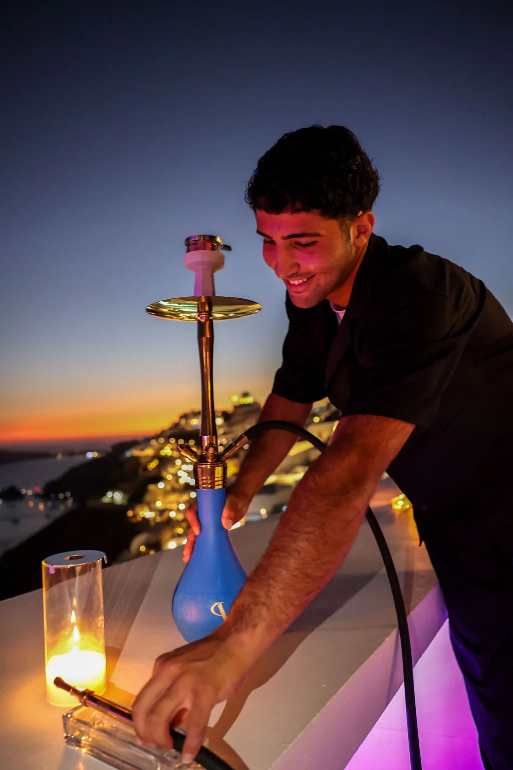 A member of our team setting a lit shisha down on the rooftop ledge at blue hour, Oia behind.