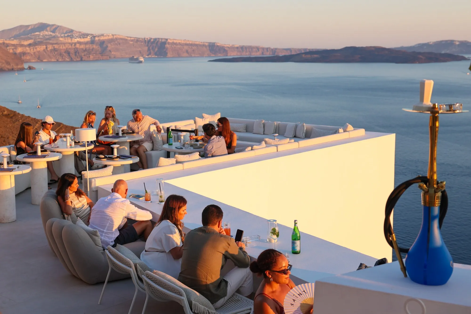 The rooftop full at golden hour, guests at the ledge and the caldera below, a shisha in the foreground.