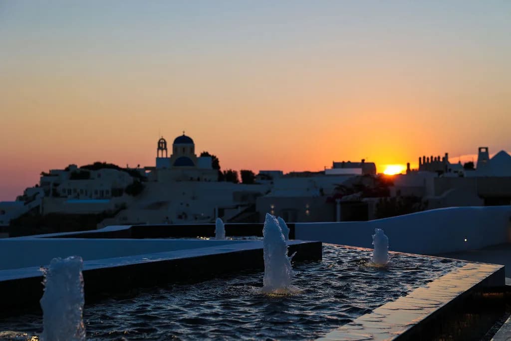 Fountain jets rising from the dark reflecting pool as the sun sets beside a blue-domed church on Oia's skyline.