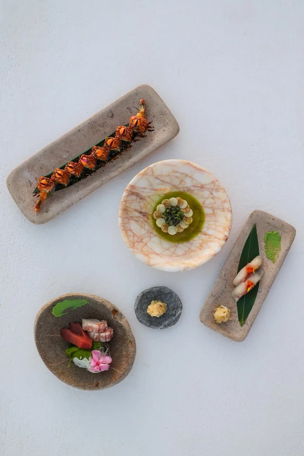 An overhead of a roll on a stone slab, a dish in green oil in a marble bowl, sashimi and two nigiri.