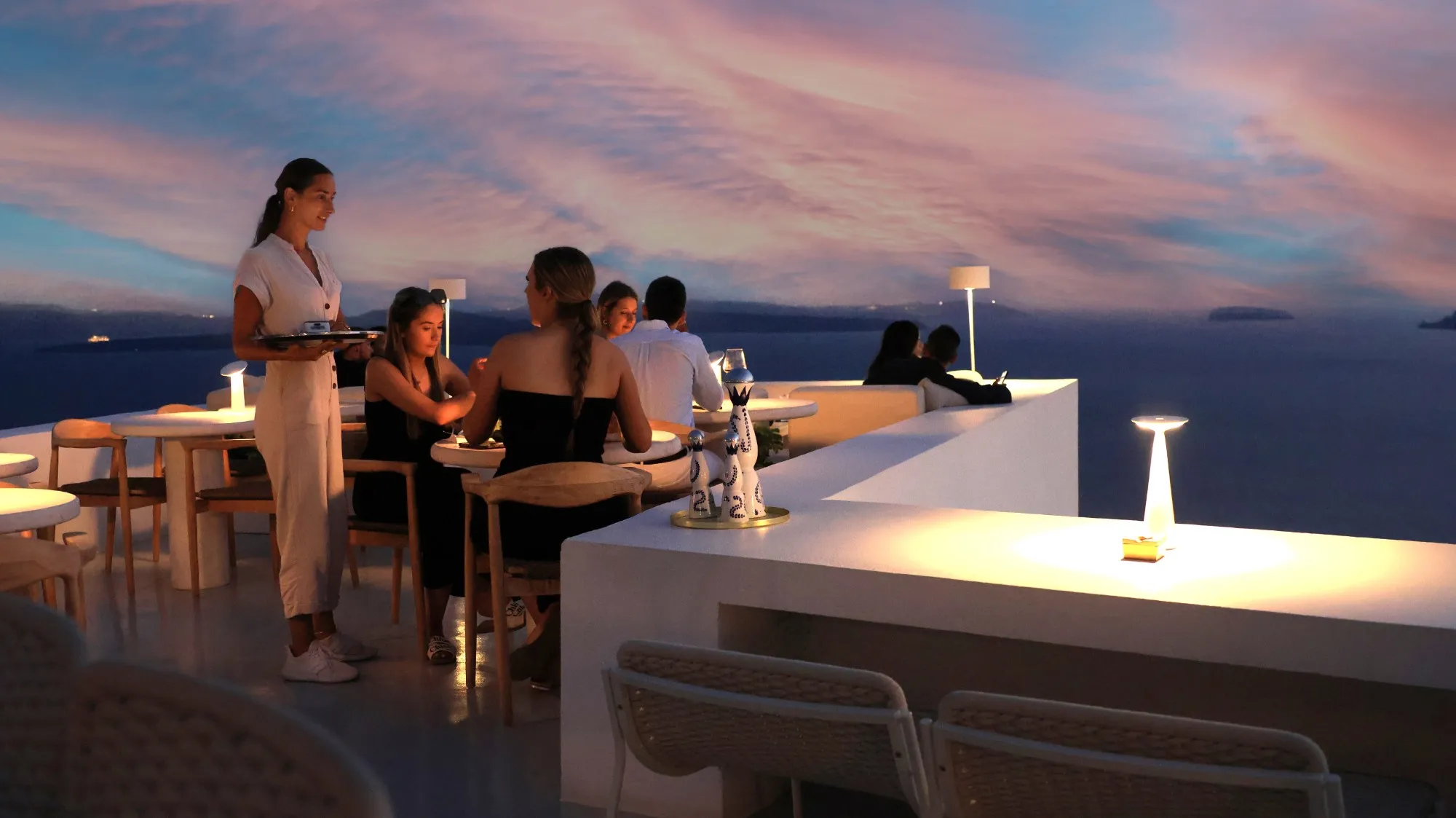 A server bringing a tray to a table of guests on the rooftop terrace as the sky turns to dusk over the sea.