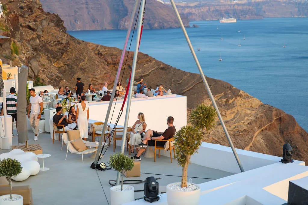 Guests in the rooftop lounge beneath the tripod's steel legs, the red cliff dropping to a caldera dotted with sailboats and a cruise ship.