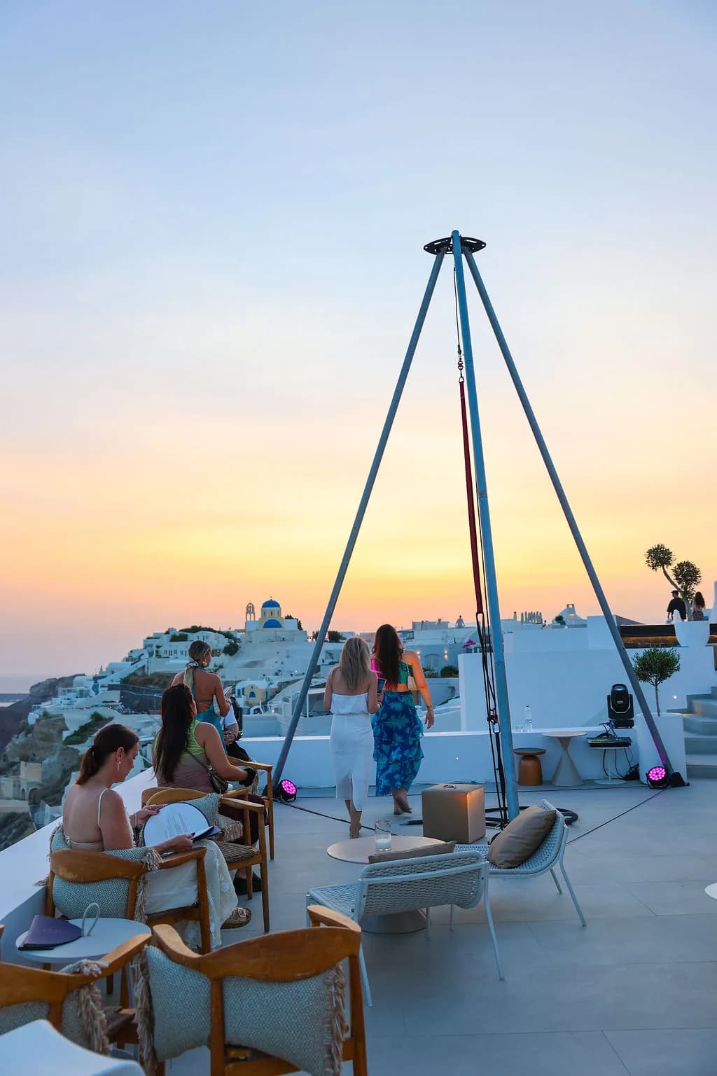 The steel tripod rig on the rooftop under a pastel sunset, guests below and Oia behind.