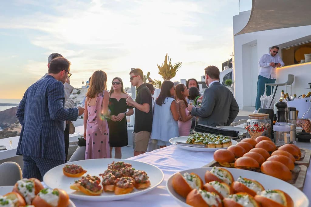 Platters of brioche sliders and tartare toasts on a white-clothed table, guests mingling on the rooftop as the sun lowers over the sea.